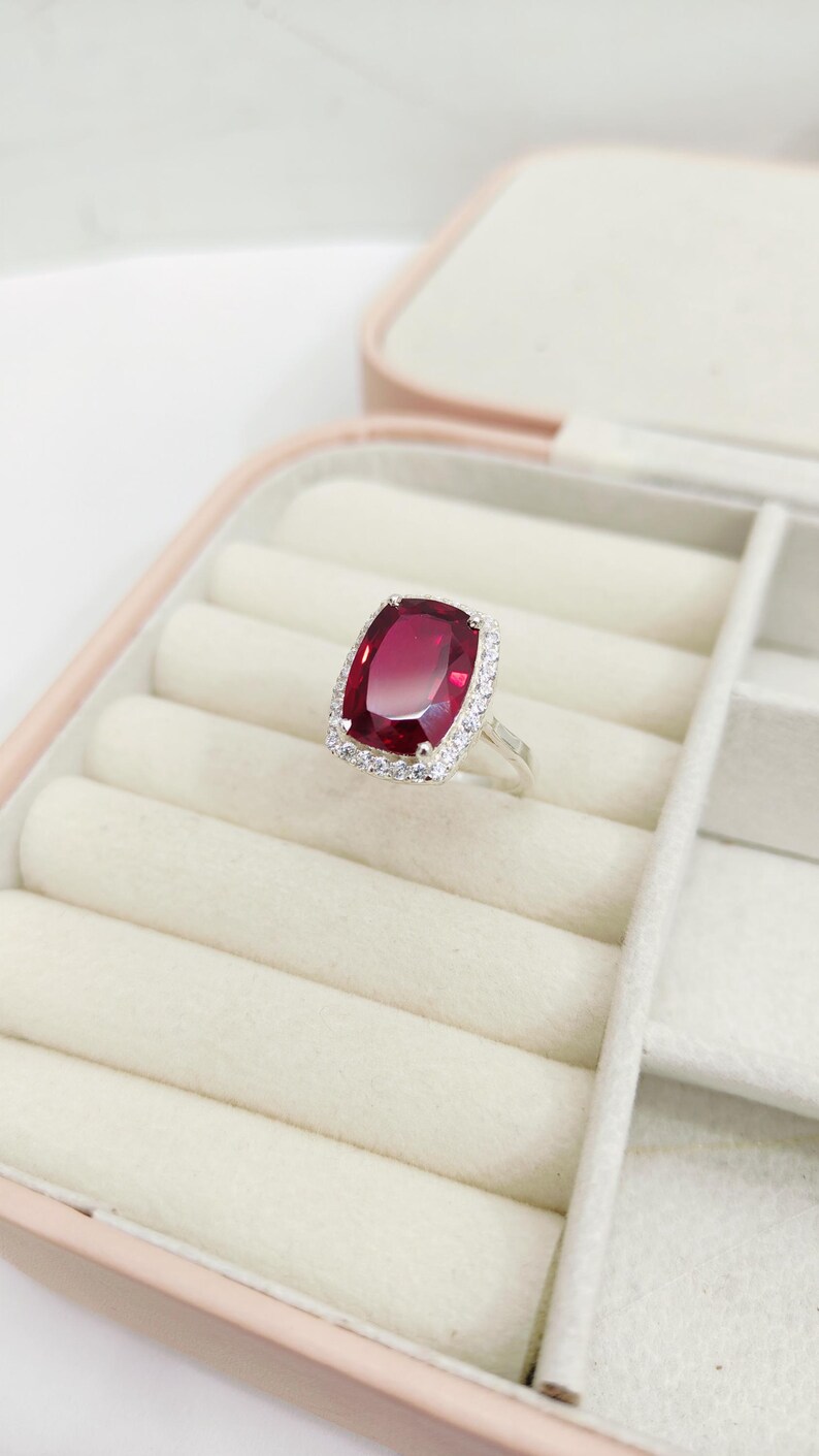 Puede incluir: Un anillo de plata con una gran gema rectangular de color rojo intenso. La gema est&aacute; rodeada por un halo de piedras m&aacute;s peque&ntilde;as y transparentes. El anillo se exhibe en un joyero rosa con interior blanco.