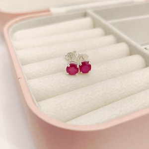 May include: A pair of round, deep pink gemstone stud earrings set in silver-toned metal prongs. The earrings feature a classic circular cut with a vibrant, saturated colour, showcasing a polished metallic finish on the posts and secure backings.