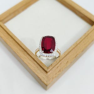 Könnte beinhalten: Ein goldener Ring mit einem großen, kissenförmigen, tiefroten Edelstein, umgeben von einem Kranz aus kleineren, klaren Steinen. Der Ring ist vor einem weißen Hintergrund in einem Holzrahmen platziert.