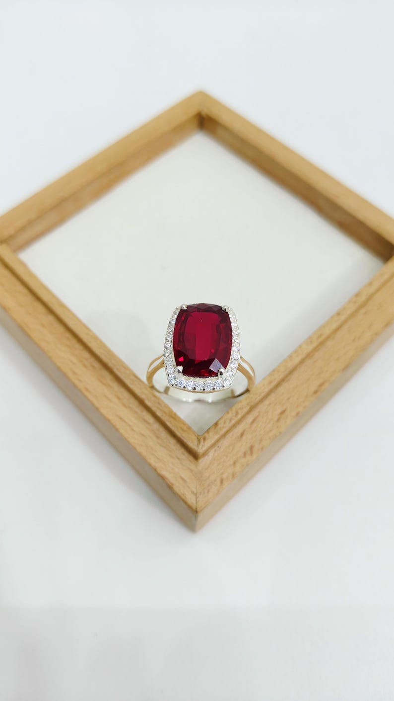 Puede incluir: Un anillo de oro con una gran piedra preciosa roja oscura de talla coj&iacute;n, rodeada por un halo de piedras m&aacute;s peque&ntilde;as y claras. El anillo est&aacute; sobre un fondo blanco dentro de un marco de madera.