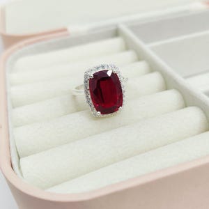 Puede incluir: Un anillo de plata con una gran gema rectangular de color rojo intenso, rodeada por un halo de piedras m&aacute;s peque&ntilde;as y claras. El anillo se exhibe en una caja de joyer&iacute;a rosa con interior blanco.