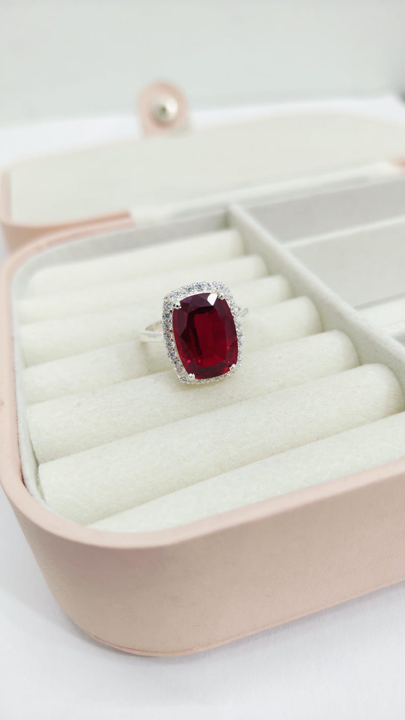 Puede incluir: Un anillo de plata con una gran gema rectangular de color rojo intenso, rodeada por un halo de piedras m&aacute;s peque&ntilde;as y claras. El anillo se exhibe en una caja de joyer&iacute;a rosa con interior blanco.