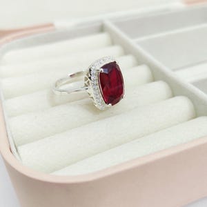 Puede incluir: Un anillo de plata con una gran gema rectangular de color rojo intenso, rodeada por un halo de piedras claras m&aacute;s peque&ntilde;as. El anillo se encuentra en un joyero rosa claro con interior blanco. La banda del anillo es delgada y pulida.