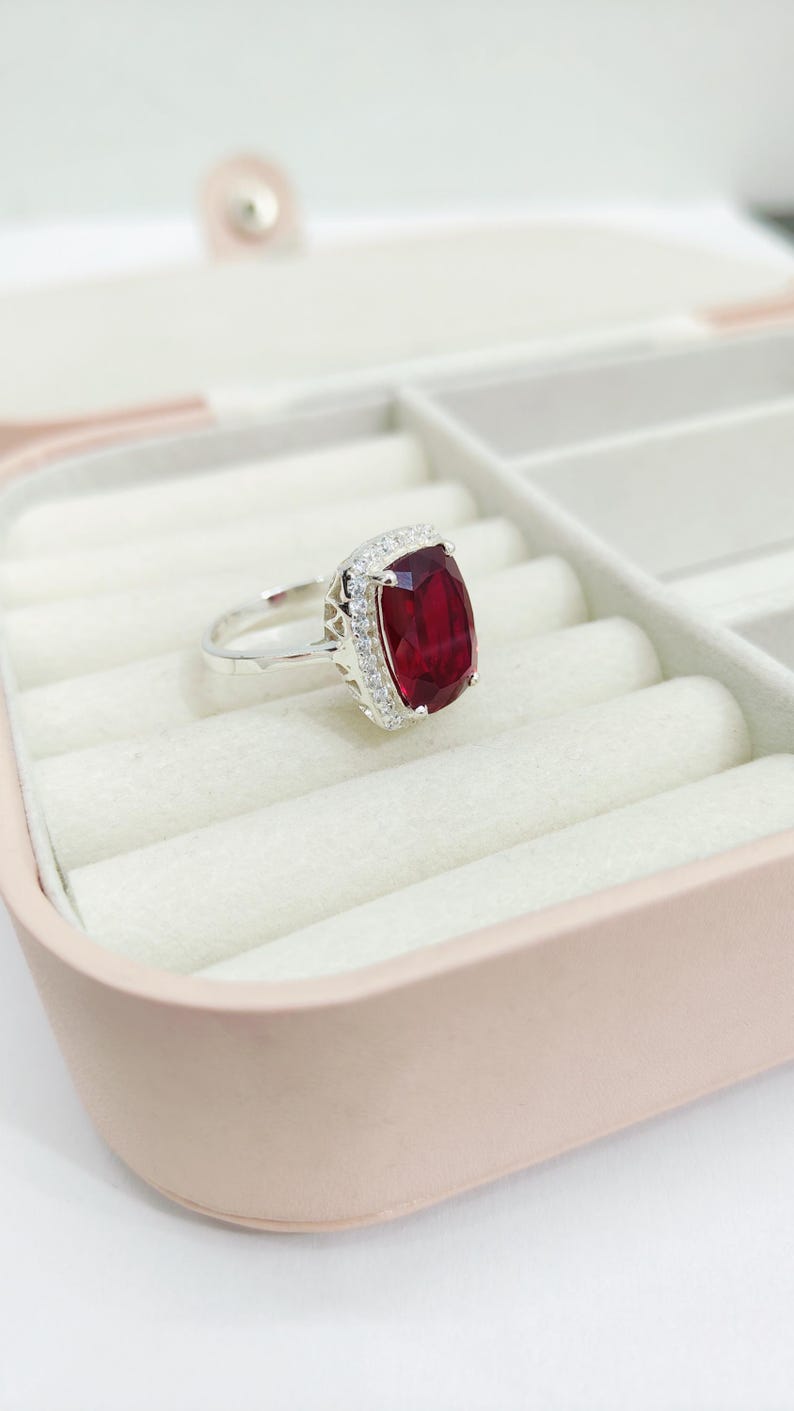 Puede incluir: Un anillo de plata con una gran gema rectangular de color rojo intenso, rodeada por un halo de piedras claras m&aacute;s peque&ntilde;as. El anillo se encuentra en un joyero rosa claro con interior blanco. La banda del anillo es delgada y pulida.