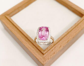 Puede incluir: Un anillo de oro con una gran gema rosa de corte cuadrado, rodeada por un halo de piedras claras más pequeñas. El anillo se presenta sobre un fondo blanco dentro de un marco de madera.