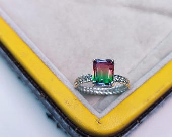 May include: A silver ring with a rectangular, multi-colored gemstone. The gemstone transitions from red to green. The ring has a twisted rope design around the band. The ring is displayed on a gray surface with a yellow border.