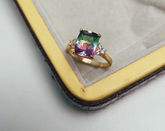 May include: A gold ring featuring a large, rectangular, multi-colored gemstone with green, purple, and pink hues. The ring is accented with smaller, clear gemstones on each side of the main stone.