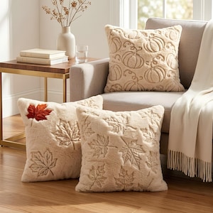Könnte beinhalten: Drei quadratische Dekokissen mit weicher, plüschiger Textur. Ein Kissen zeigt ein geprägtes Kürbismuster in Creme. Zwei Kissen zeigen geprägte Ahornblattmuster in Creme, wobei ein Blatt mit einem leuchtenden Farbverlauf in Orange und Rot akzentuiert ist.