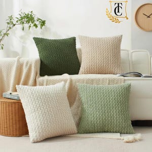 May include: Four square throw cushions with a textured, woven popcorn-style pattern. The set includes one dark forest green, one beige, one cream, and one sage green cushion, all featuring a consistent raised knit design on the front surface.