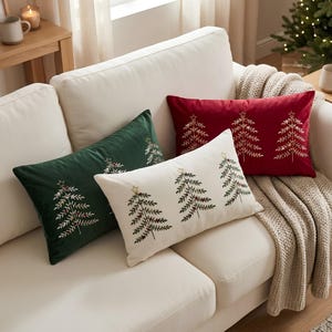 May include: Three rectangular throw pillows in dark green, cream, and deep red velvet. Each pillow features embroidered gold-toned Christmas tree designs with star toppers and small red accents, arranged in a row on a light-colored sofa.