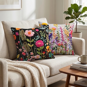 Può includere: Due cuscini decorativi con disegni floreali. Un cuscino ha uno sfondo nero con fiori rosa, arancioni e gialli. L'altro cuscino ha uno sfondo bianco con fiori colorati. I cuscini sono su un divano beige.