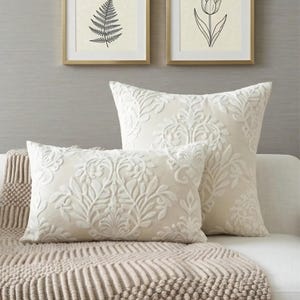 Könnte beinhalten: Zwei cremefarbene Dekokissen mit einem erhabenen, Ton-in-Ton gehaltenen Damast-Blumenmuster. Ein Kissen ist quadratisch, das andere rechteckig; beide zeigen eine komplexe, symmetrische florale Stickerei auf einem glatten, einfarbigen Stoff.