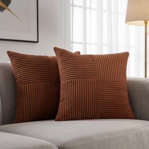 May include: Two square, rust-colored throw pillows with a corduroy texture rest on a light gray sofa. The pillows have a patchwork design, with vertical and horizontal ribbed sections. The pillows add a touch of warmth and texture to the neutral-toned living space.