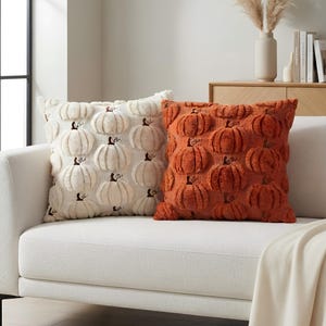 May include: Two square throw pillows featuring a raised, textured pumpkin pattern. One pillow is cream-colored, and the other is a burnt orange shade. Both pillows have a soft, plush surface with a repeating design of pumpkins across the front.
