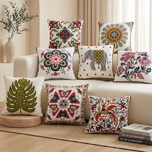 May include: A collection of eight square throw pillows featuring diverse embroidered designs including a green monstera leaf, a colorful elephant, floral patterns, butterflies, and a bird, all displayed on a light-colored sofa with textured fabric.