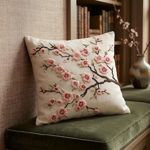 Può includere: Cuscino quadrato color crema con un ricamo di fiori di ciliegio. Il design presenta fiori rosa, rami marroni e foglie verdi. Il cuscino è posto su una superficie di velluto verde. Il testo "Sakura Atelier" è visibile.