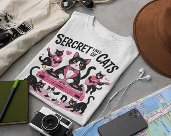 May include: White t-shirt with the text "Secret Lives of Cats" and a graphic of cats playing instruments and a DJ cat. Other items include a camera, notebook, sunglasses, and a brown hat.