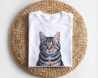 May include: White t-shirt with a detailed illustration of a grey tabby cat. The cat has green eyes and is centered on the shirt. The shirt is folded and placed on a round, woven, light brown mat.