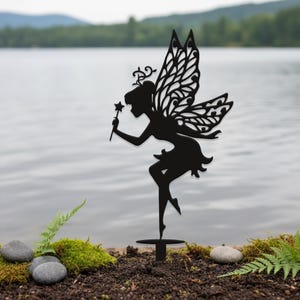 Könnte beinhalten: Schwarze Metall-Feengartenstecker. Die Silhouette zeigt eine Fee mit detaillierten Flügeln, die einen sternförmigen Zauberstab hält. Die Fee ist in einer anmutigen Pose mit fließendem Rock dargestellt. Der Stecker ist zum Einsetzen in den Boden konzipiert.