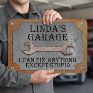 Könnte beinhalten: Rechteckiges Metallschild mit dem Text "LINDA'S GARAGE" über einer Schraubenschlüssel-Grafik und dem Satz "I CAN FIX ANYTHING EXCEPT STUPID". Das Schild hat einen rostbraunen Rand und wird von einer Person gehalten.