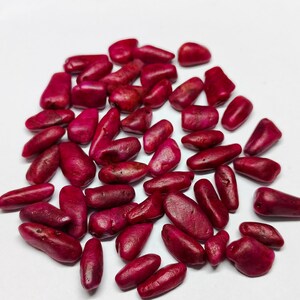 May include: A collection of polished, deep red gemstones, possibly coral or beads, scattered on a white surface. The stones vary in size and shape, with a smooth, glossy texture. The colour is a rich, saturated red.
