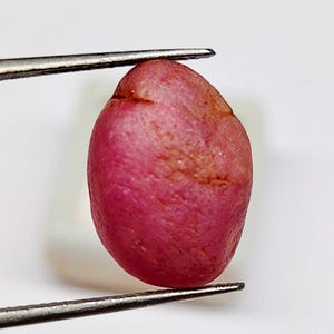 May include: A raw, unpolished gemstone with a rounded, oval shape and a vibrant pinkish-red hue. The surface of the stone displays a natural, textured appearance with subtle variations in colour intensity and small surface irregularities.