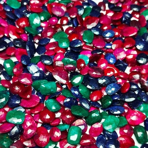 May include: A collection of faceted gemstones in various colors, including red, green, and blue. The gemstones are scattered, showcasing their different shapes and sizes. The image is well-lit, highlighting the sparkle and brilliance of the stones.