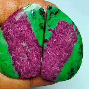 May include: Two polished gemstone cabochons. Each stone has a teardrop shape with a vibrant green border and a central area of deep magenta with a fibrous texture. The stones are held in a hand.
