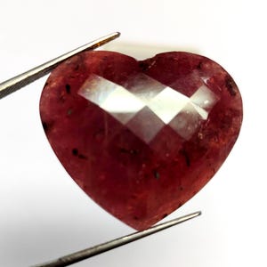 May include: A heart-shaped, faceted gemstone in shades of red, held by tweezers. The gem has a complex, multi-faceted surface that reflects light. Small dark inclusions are visible within the stone, adding to its unique appearance.