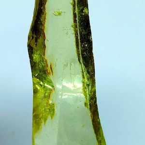 Puede incluir: Un fragmento translúcido y alargado de vidrio color verde lima con bordes irregulares y dentados, y una superficie lisa y reflectante. La pieza muestra tonos variables de verde amarillento y secciones claras con burbujas internas visibles.
