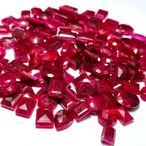 Könnte beinhalten: Eine Ansammlung facettierter, tiefroter Edelsteine, darunter Rubine, in verschiedenen Formen wie oval, rechteckig und quadratisch. Die Edelsteine liegen auf einer weißen Oberfläche und zeigen ihre Brillanz und satte Farbe.