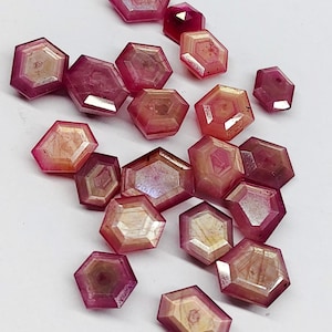May include: A collection of loose, faceted gemstones in a hexagonal shape. Each stone features a gradient of deep magenta and pink hues transitioning into a pale, cream-colored center, showcasing a distinct geometric pattern and polished surface.