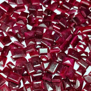 Puede incluir: Un conjunto de gemas facetadas de color rojo intenso en varias formas, incluyendo rectángulos, cuadrados y óvalos. Las gemas están dispersas sobre una superficie blanca, mostrando sus superficies pulidas y su rico color.