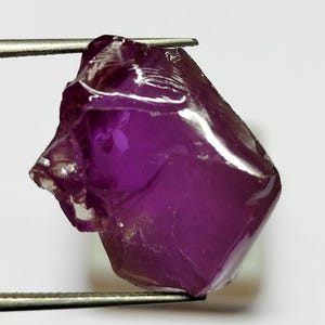 Puede incluir: Una piedra preciosa en bruto de color púrpura intenso con una estructura cristalina translúcida y bordes irregulares y facetados. La piedra está sujeta por unas pinzas metálicas delgadas de tono plateado, mostrando su color vibrante y textura natural.