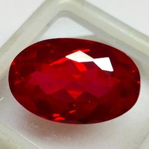 Peut inclure: Une pierre précieuse rouge foncé, de forme ovale et à facettes. La gemme est placée dans un support en plastique transparent, et les facettes réfléchissent la lumière, créant des reflets brillants. L'aspect général est vif et poli.