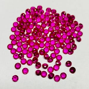 Puede incluir: Una colección de pequeñas gemas redondas, facetadas, de color rosa intenso, dispersas sobre una superficie blanca. Las gemas tienen un corte brillante, que refleja la luz. Las gemas son de un color vibrante y saturado, y parecen ser rubíes o piedras preciosas similares.