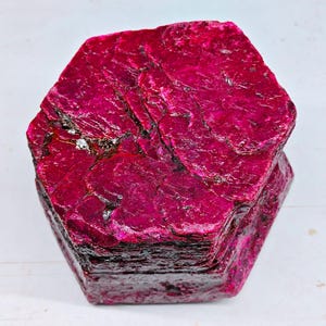 Puede incluir: Un cristal de rubí hexagonal en bruto con una paleta de colores magenta y carmesí intenso. La superficie muestra un aspecto natural y texturizado con fisuras visibles, capas cristalinas e inclusiones oscuras en la estructura.