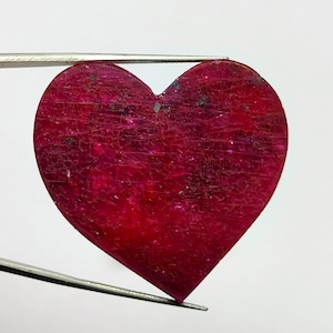 May include: A heart-shaped, deep red gemstone with a textured surface. The gemstone is held by a pair of silver tweezers against a white background. The stone has a slightly irregular shape and a polished appearance.