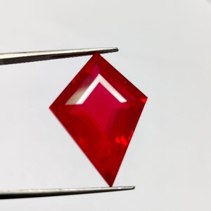 May include: A faceted, red gemstone held by tweezers. The gem is diamond-shaped with a white, triangular facet at the top. The gem's color is a vibrant, deep red, and it appears to be highly polished.