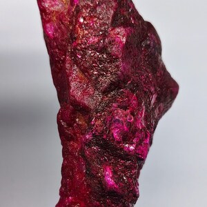 Peut inclure: Une pierre précieuse rubis brute et non polie d'une couleur magenta et cramoisi profonde et vibrante. La pierre présente une surface irrégulière et très texturée avec des facettes cristallines naturelles et des inclusions sombres.