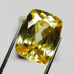May include: A large, faceted yellow gemstone held by tweezers. The gem has a rectangular shape with rounded corners and a complex internal structure, reflecting light in various directions. The color is a vibrant yellow with hints of gold and white.