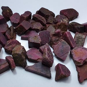 May include: A pile of raw ruby gemstones in shades of deep red and purple. The stones vary in size and shape, with some having rough, textured surfaces and others smoother facets. The gemstones are scattered on a white surface, showcasing their natural beauty.