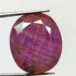 May include: Oval-shaped, faceted ruby gemstone held by tweezers. The ruby displays a deep pinkish-red hue with internal striations and reflective facets. The gemstone is a polished, natural mineral, suitable for jewelry making.