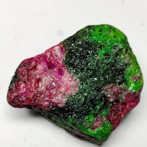 Peut inclure: Une pierre précieuse brute avec un mélange de couleurs vives. La pierre présente des teintes rouge foncé et magenta, contrastant avec des zones de vert et de noir riches. La texture naturelle et les variations de couleur créent un aspect unique.