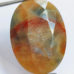 May include: Oval-shaped gemstone with a faceted surface. The gem displays a blend of colors, including orange, yellow, red, and hints of green. Held by a pair of silver tweezers, the stone's polished facets catch the light, showcasing its natural patterns.