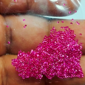 May include: A close-up of a pile of small, bright pink gemstones. The gemstones are scattered across a surface, with some loose gems around the main pile. The gems are sparkling and appear to be faceted.