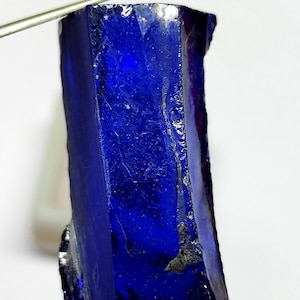 May include: A raw, deep blue sapphire crystal with a columnar, elongated shape. The gemstone features a translucent to opaque appearance with natural surface textures, jagged edges, and visible internal inclusions throughout the stone.