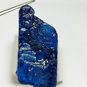 May include: A deep blue, raw sapphire gemstone with a rough, textured surface. The sapphire is held by tweezers, showcasing its irregular shape and natural beauty. The gemstone displays varying shades of blue.