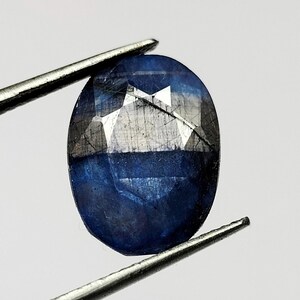 Puede incluir: Una gema de zafiro ovalada y facetada, sujeta con pinzas. El zafiro muestra un color azul profundo con facetas más claras y reflectantes. La gema se encuentra sobre un fondo blanco liso, resaltando su claridad y brillo. Una vista de cerca muestra el intrincado corte de la gema.