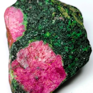 Könnte beinhalten: Ein roher Edelstein mit leuchtend rosa Rubinkristallen, eingebettet in eine Matrix aus dunkelgrünen und schwarzen Mineralien. Der Stein hat eine strukturierte Oberfläche mit Bereichen in leuchtenden Grün- und Rosatönen.